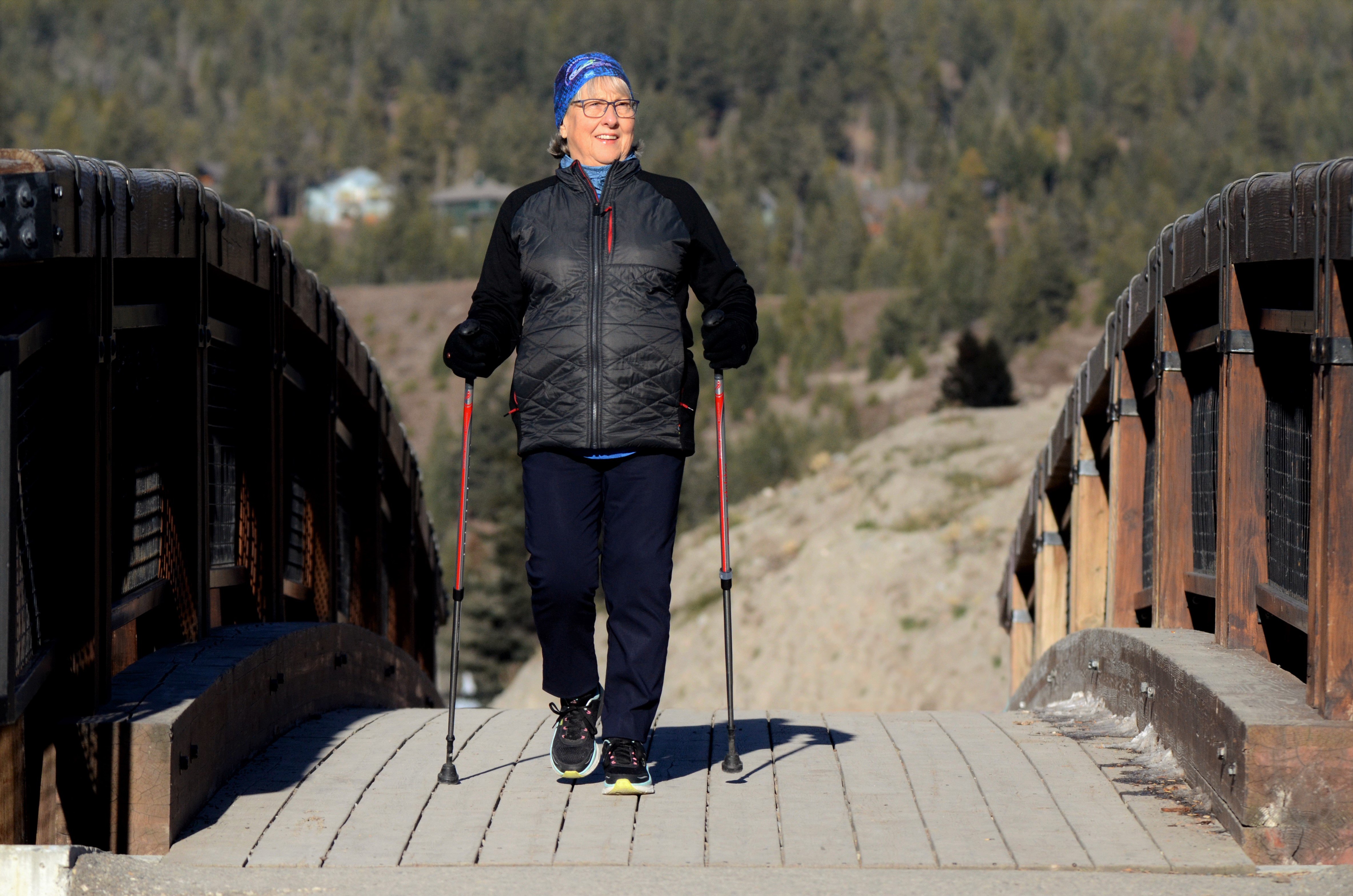 Marge with her Origin PowerStride polls walking across a wooden bridget with a mountain side and trees in the background