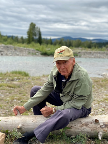 Yvan sitting on a log enjoying the great outdoors beside a lake and mountains in the background